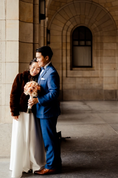 A loving and intimate moment of the bride and groom outside the gemeente Rotterdam after their wedding ceremony. The bride and groom share a tender embrace, with the grand architecture of gemeente huis as the backdrop. The bride rests her head on the groom's shoulder as light streams in, creating a romantic and visually stunning wedding photo.