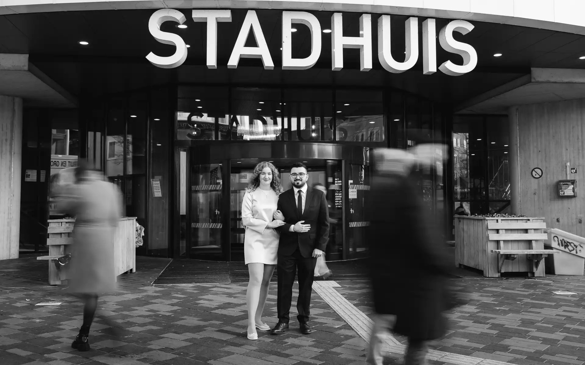 This modern black and white wedding photo captures the joy and intimacy of an Almere couple in front of the Stadhuis. The bride and groom are looking directly into the camera, their eyes filled with happiness and love. The blurred background, with people passing by, creates a sense of privacy and isolation, as if the couple is the only one in the world on their special day – captured by DiscussTomorrow Studio.