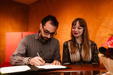 The joyous moment of signing the marriage deeds captured by DiscussTomorrow Studio. The bride's radiant smile lights up the warm and luxurious room at gemeente Almere’s Trouwzaal as she watches her husband sign, creating a beautiful and intimate memory of their special day.